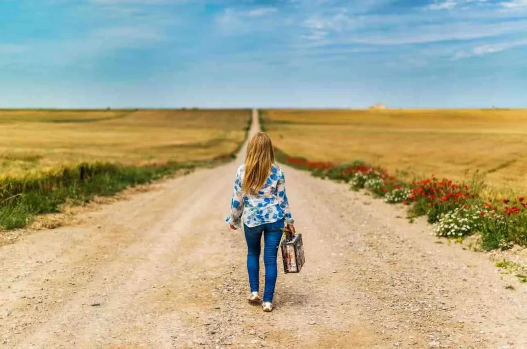 C'est une femme qui avance sur un chemin de terre avec une valise à la main