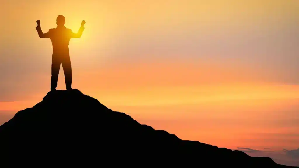 Une personne est au sommet d'une montagne avec les mains vers le ciel. Le ciel est orangé.